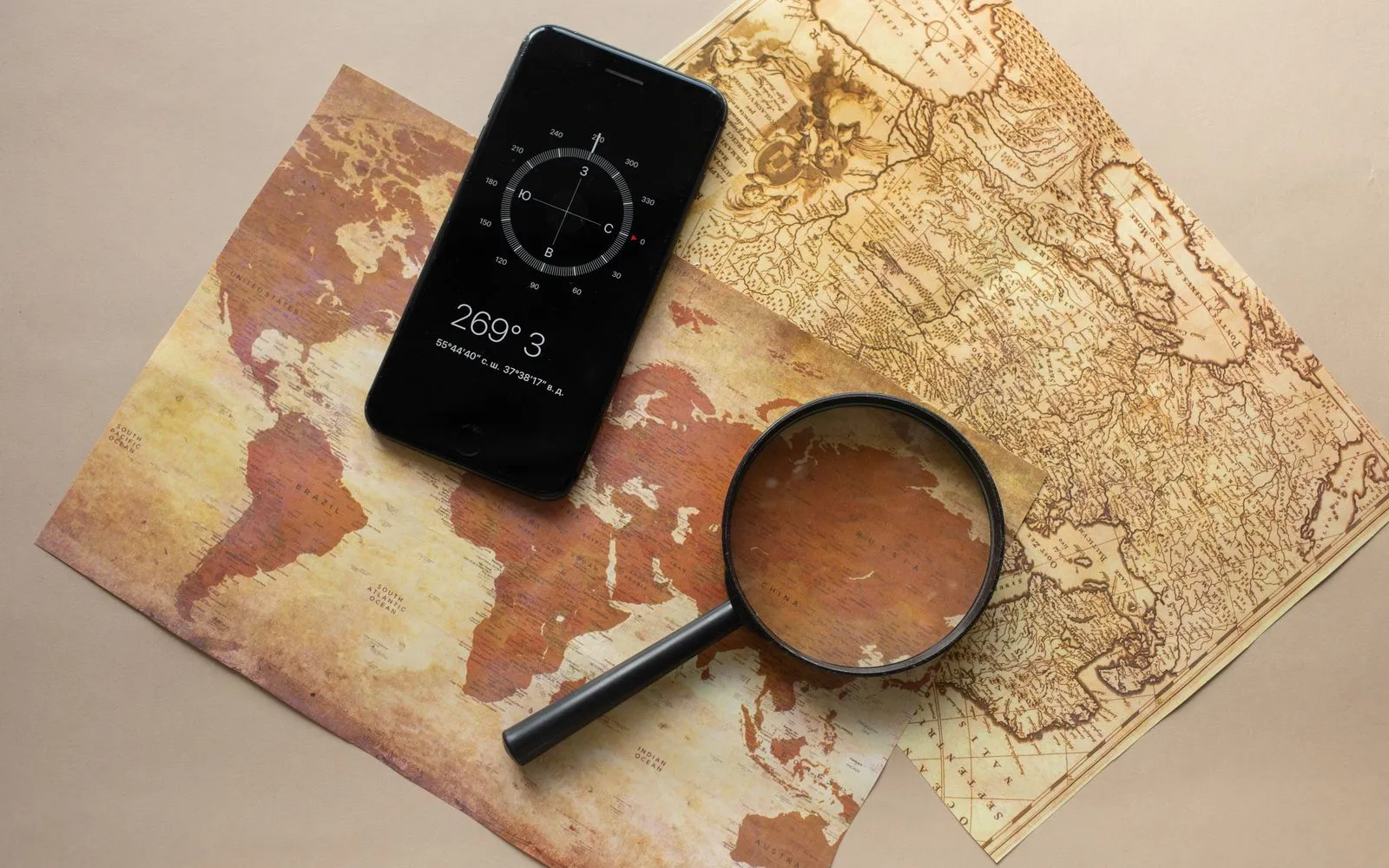 Top view of magnifying glass and cellphone with compass with coordinates placed on paper maps on beige background in light room